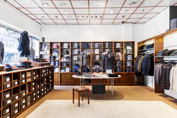 Intérieur d'une boutique de mode bien organisée avec des étagères en bois remplies de vêtements, chemises, pantalons et accessoires soigneusement présentés. Un comptoir central avec un tabouret pour accueillir les clients.